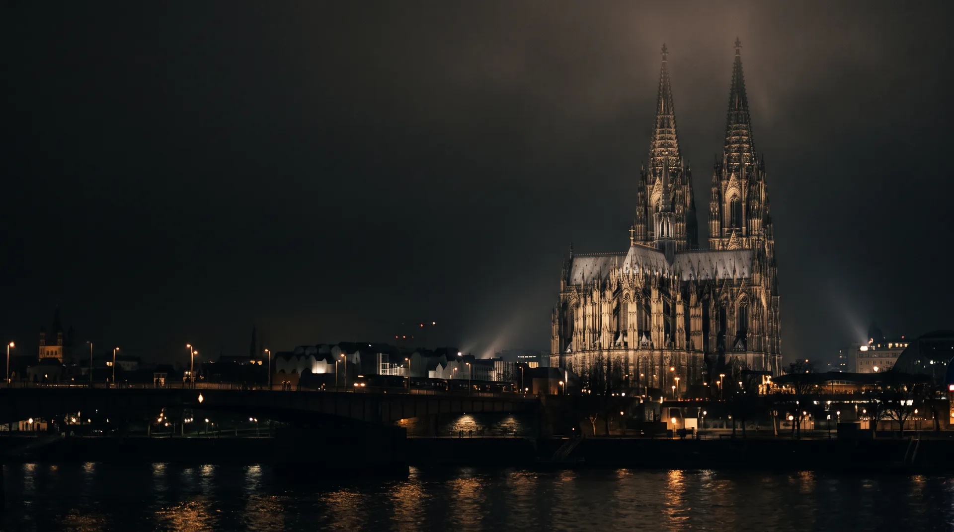 Kölner Dom bei Nacht in warmem Licht, davor der dunkle Rhein
